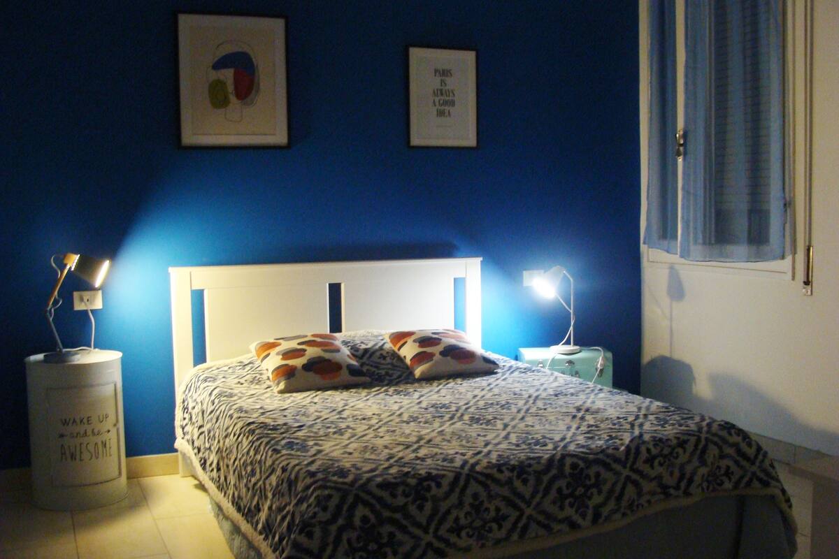 Une chambre à coucher avec une couvre-lit à motifs, deux oreillers, une table de chevet avec une lampe et un mur orné de deux tableaux encadrés.