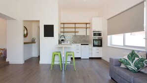 A modern kitchen with white cabinets, a built-in oven, and a dining area with two green stools. - Koala at White Gum Lodge (White Gum Valley)