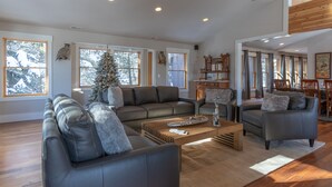 A spacious living room with a large sectional sofa, a coffee table, and a Christmas tree. There are large windows with curtains, a wooden ceiling, and a dining area with chairs and a table. - Trinity :Sleeps 10- HotTub-GameRoom-Huge Kitchen-Perfect4Groups Tram & Mtn View (Teton Village)