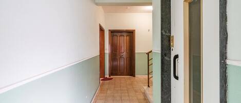 A narrow hallway with a wooden door at the end, a handrail on the right, and a tiled floor.