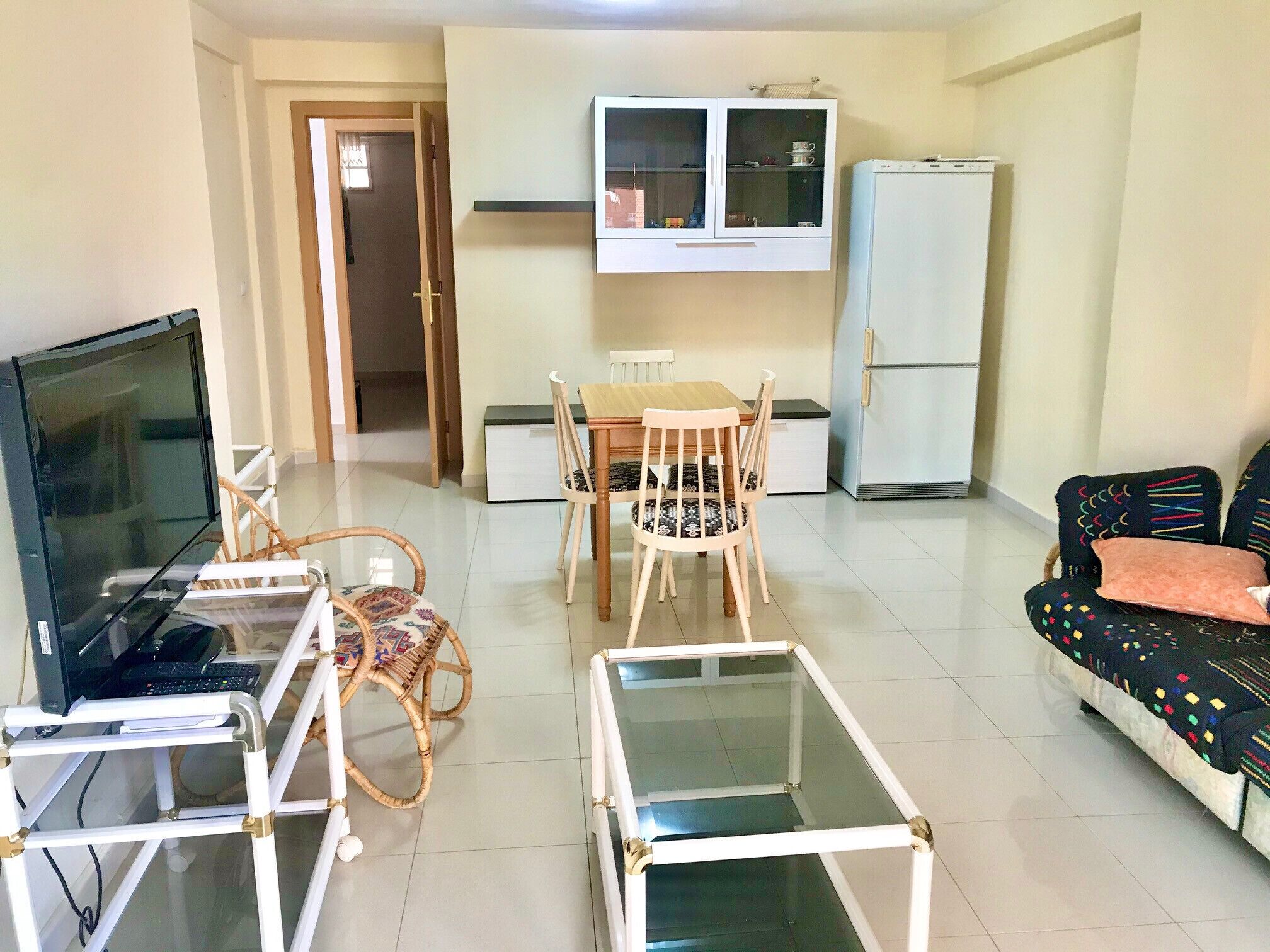 A living room with a TV, a glass coffee table, a sofa, a dining table with chairs, a cabinet, and a refrigerator.