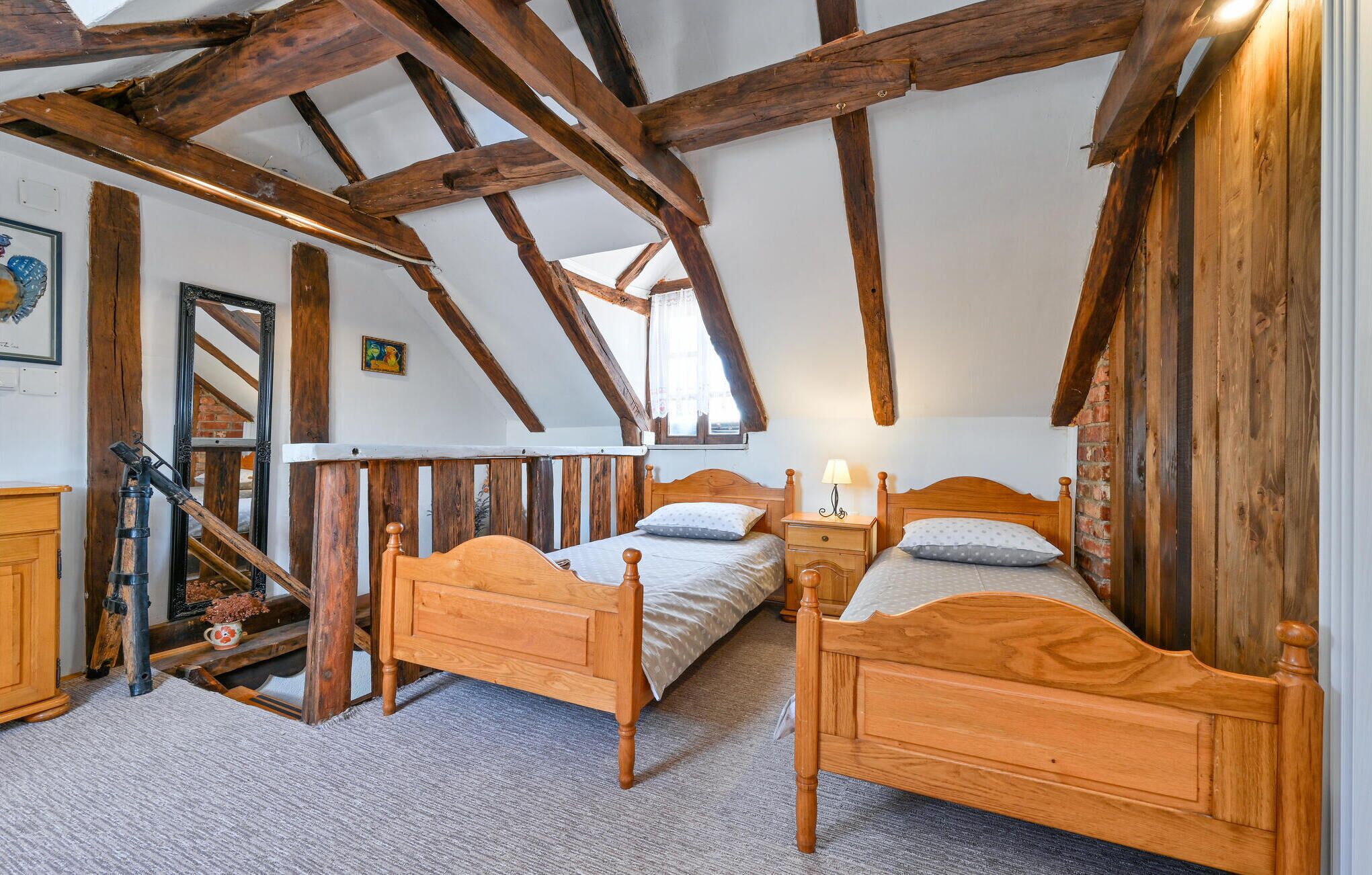 A rustic bedroom with exposed wooden beams, two wooden beds, a small bedside table with a lamp, and a mirror on the wall.