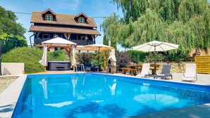 Una piscina con lettini e zona barbecue, circondata da un pontile in legno e un'ampia casa con tetto in tegole.