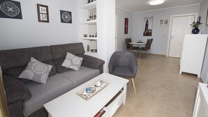 Ein modernes Wohnzimmer mit einem grauen Sofa, einem weißen Couchtisch und einem grauen Sessel. Im Hintergrund befindet sich ein Essbereich mit Tisch und Stühlen.