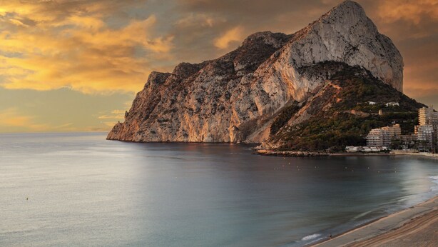 Paesaggio costiero con una montagna rocciosa imponente, mare calmo e alcuni edifici sulla riva.