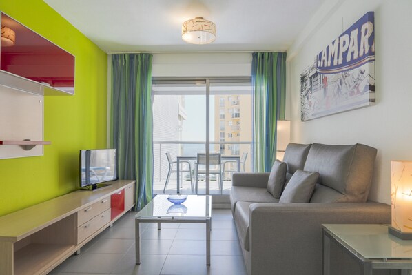 Een moderne woonkamer met een grijze bank, een glazen salontafel, een flatscreen televisie op een houten kast en een balkon met groene gordijnen.