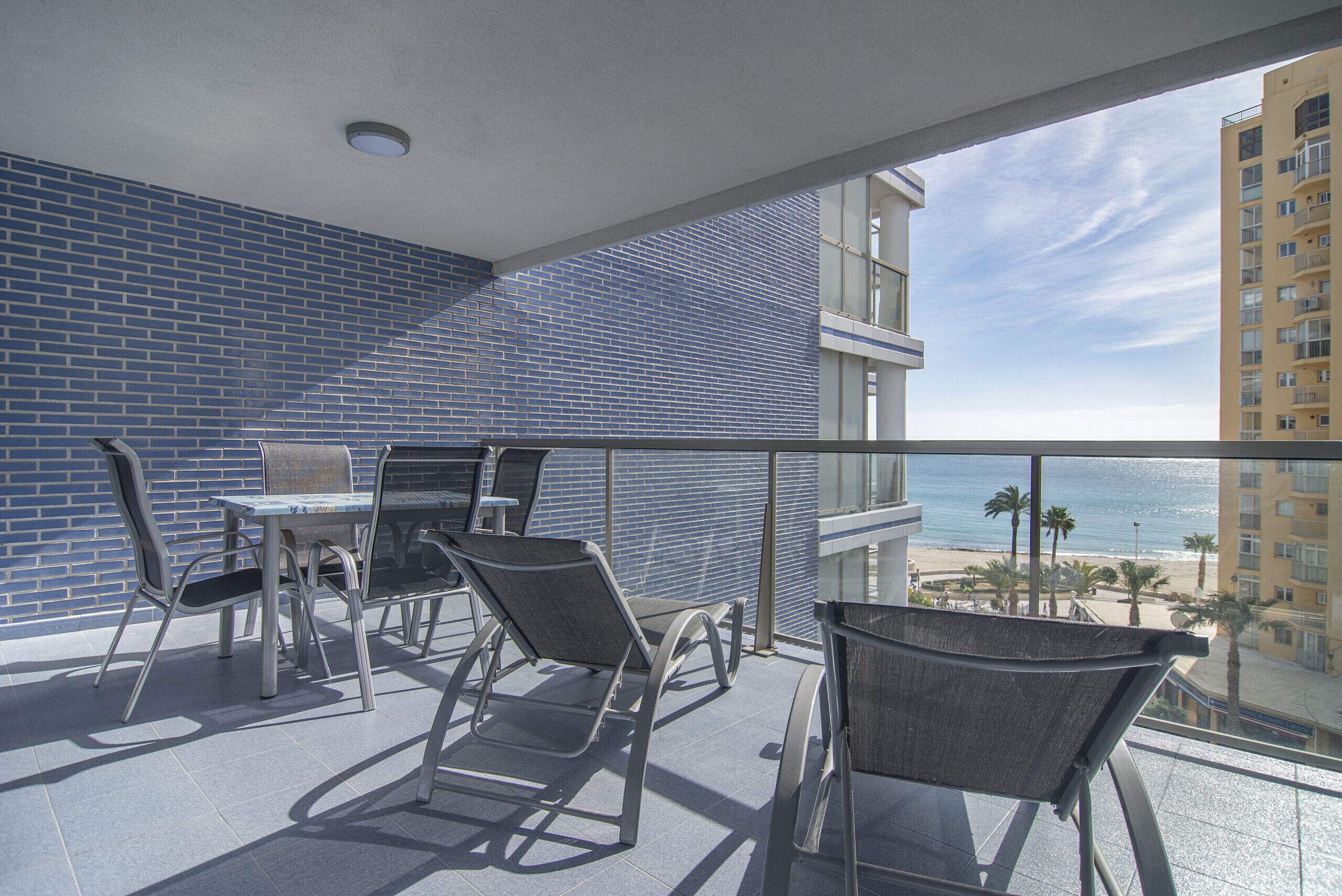 A balcony with a table and chairs, a view of the beach, and a tall building in the background.