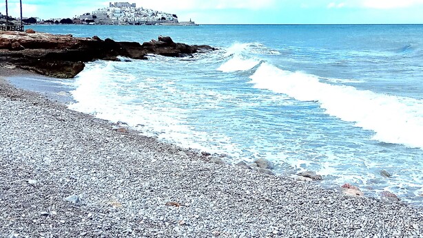 A pebble beach with waves crashing onto the shore, a rocky outcrop, and a distant building on a hill. - Edison Bungalow LEK (Peñiscola)