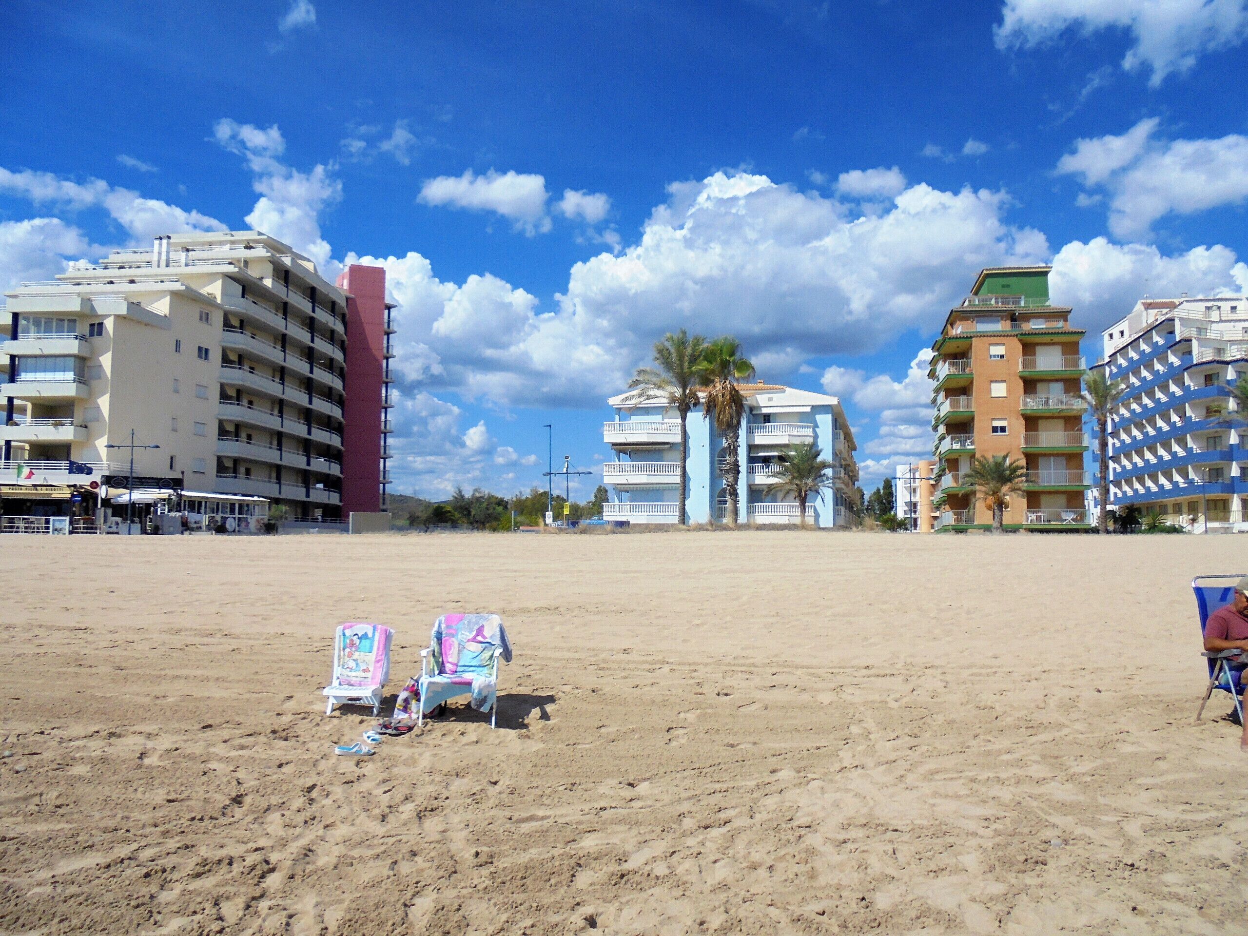 A beach with a row of multi-story buildings, some with balconies, and a few palm trees.