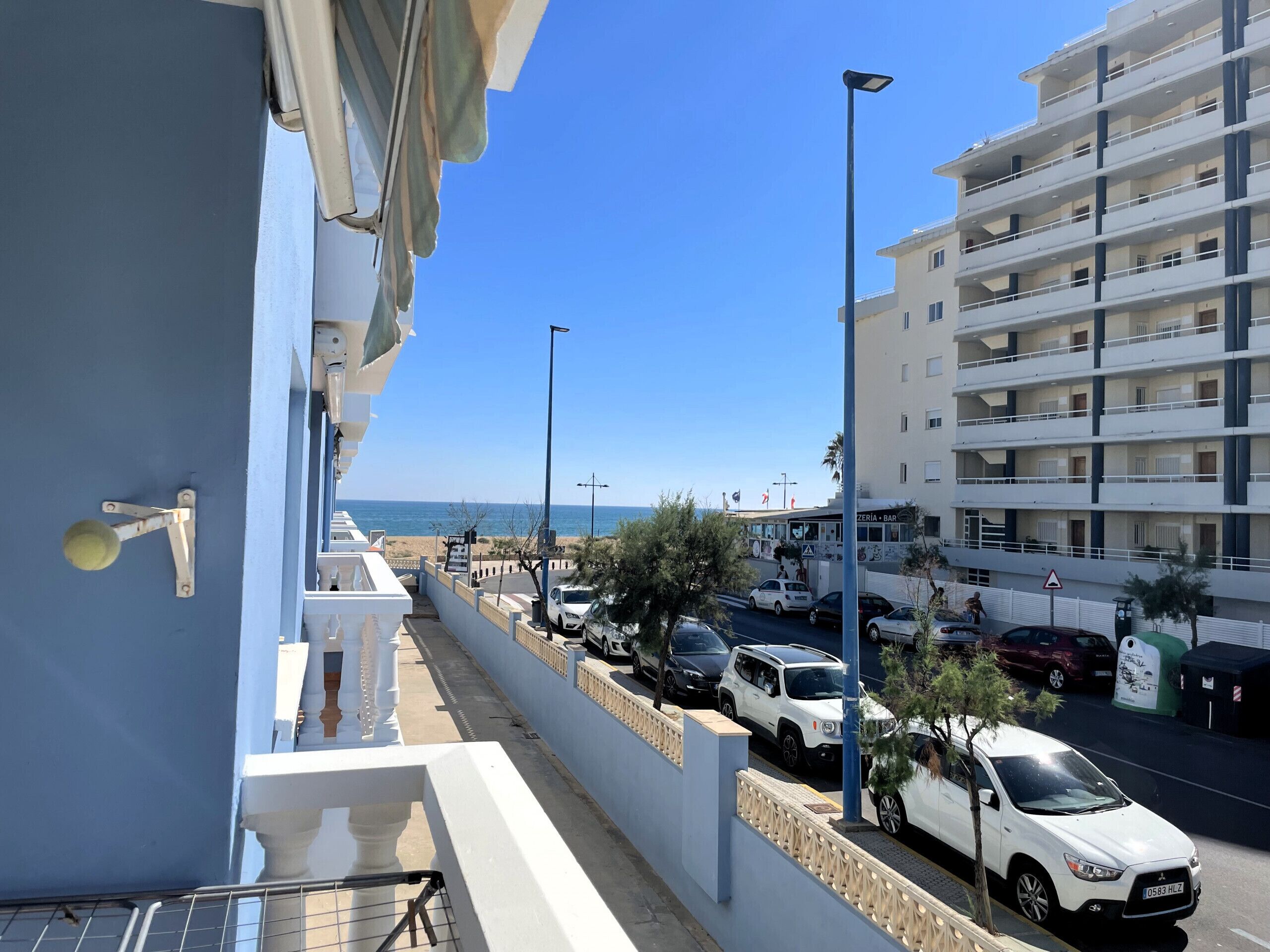 A coastal street with parked cars, a building with balconies, and a clear view of the sea.