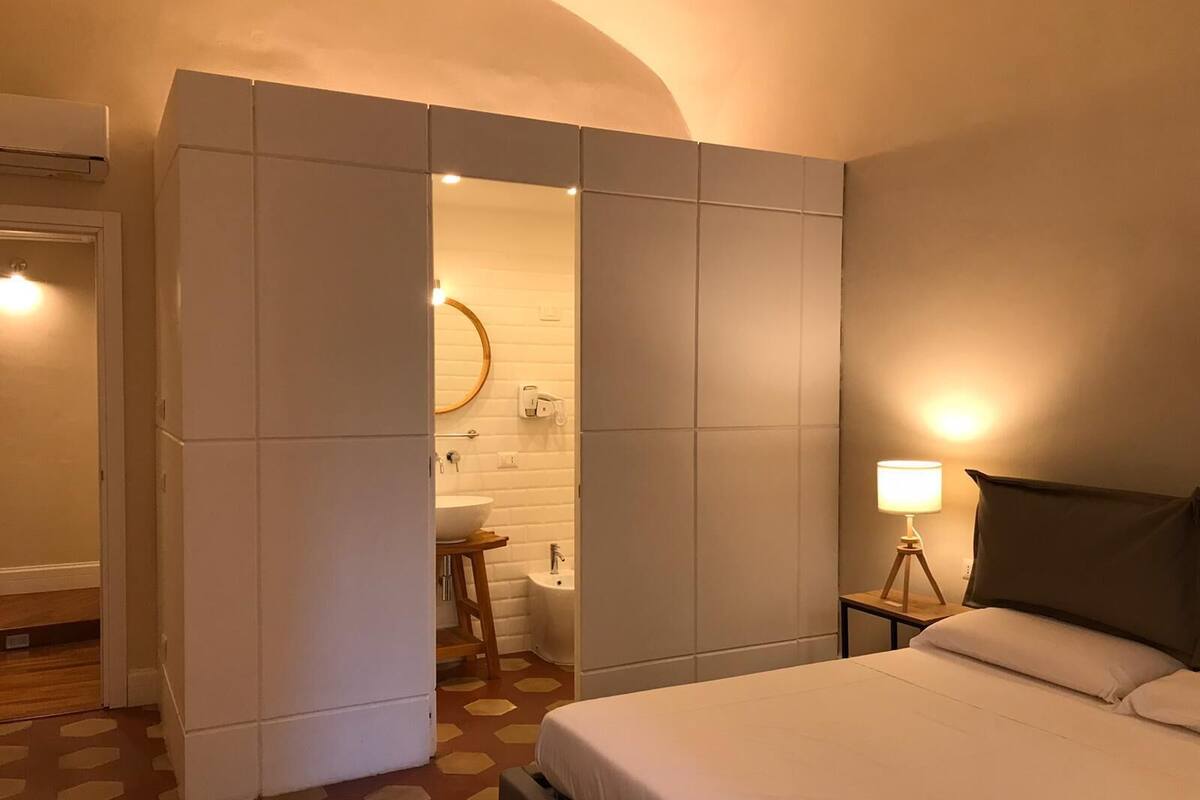 A hotel room with a bed, a bedside lamp, a bathroom with a sink and toilet, and a wardrobe.