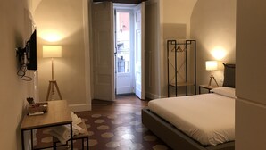 Camera d'albergo con un letto, una scrivania con una lampada, una TV a parete e vista sull'esterno attraverso una porta aperta.