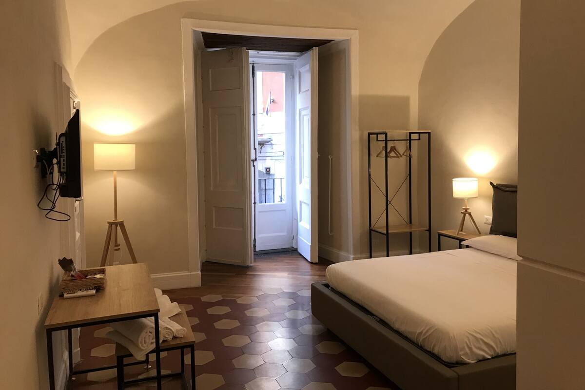 A hotel room with a bed, a desk with a lamp, a TV mounted on the wall, and a view of the outside through an open door.