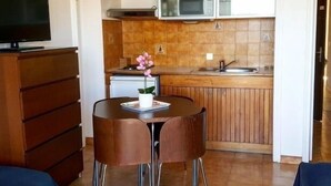 A compact kitchen with a microwave, sink, and wooden cabinets. There is a dining area with a table and chairs, and a television on a dresser. - Studio Orsoni - See View (Calvi)