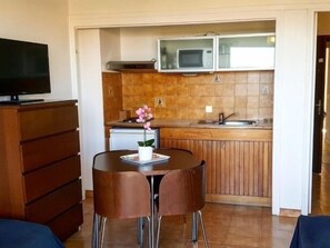 Ett kompakt kök med mikrovågsugn, diskho och träskåp. Det finns en matplats med ett bord och stolar, och en tv-apparat placerad på en byrå.