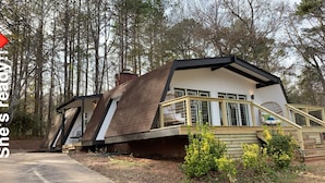 Una casa con techo inclinado, un porche con barandilla y una ventana que ofrece vistas del bosque circundante.
