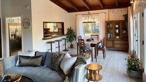 Un salón acogedor con un sofá gris, vigas de madera en el techo, una zona de cocina con nevera y una mesa de comedor con sillas.