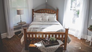 A neatly made bed with a wooden headboard, a bedside table with a lamp, and a window with sheer curtains. - Close to everything! DISCOUNTS for Long Stays- Fully Remodeled (Eau Claire)