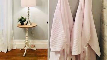 A bathroom with a wooden floor, a white wall, a small round table with a lamp, and two towels hanging on a black hook.