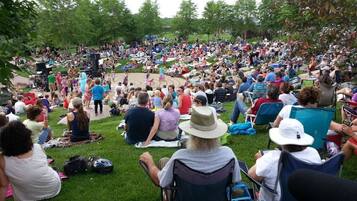 A large outdoor gathering with people seated on grass, some on chairs, and a stage with musical equipment in the background.