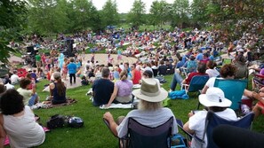 A large outdoor gathering with people seated on grass, some on chairs, and a stage with musical equipment in the background.