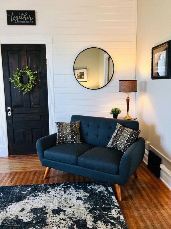A dark blue sofa with patterned pillows, a round mirror, a small plant, and a black and white rug in a room with wooden flooring and a dark door.