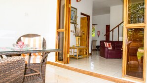 A dining area with wicker furniture, a glass table, and a view into a living room with a sofa and a staircase.
