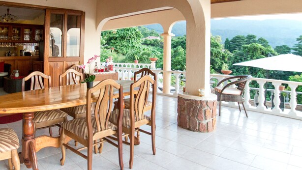 A covered dining area with a wooden table and chairs, a bar area with shelves, and a view of lush greenery.