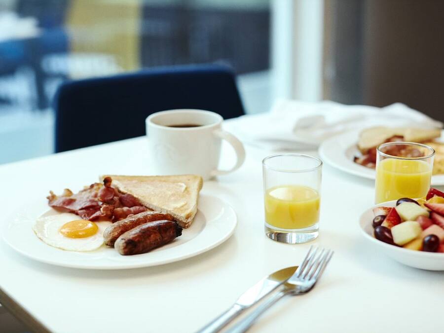 A breakfast table with toast, eggs, sausages, bacon, coffee, orange juice, and a fruit bowl.