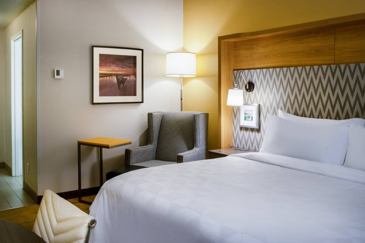 Een hotelkamer met een bed, een stoel, een klein tafeltje, een lamp en een foto aan de muur.
