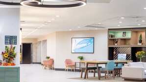 Un moderno vestíbulo de hotel con mostrador de recepción, señalización direccional, un área de comedor con sillas, un sofá y una gran obra de arte enmarcada en la pared.