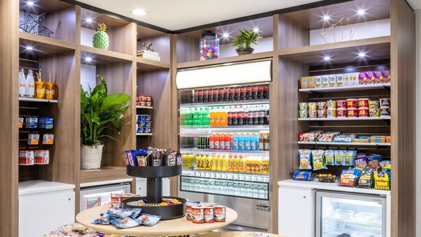 Una sección bien organizada de aperitivos y bebidas, con estantes llenos de diversos productos, un refrigerador expositor y una pequeña mesa con snacks.