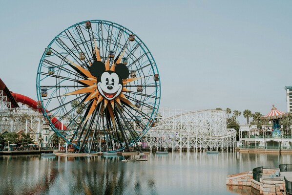 Una noria grande con diseño de la cara de Mickey Mouse, rodeada de agua y otras atracciones de parque de diversiones.