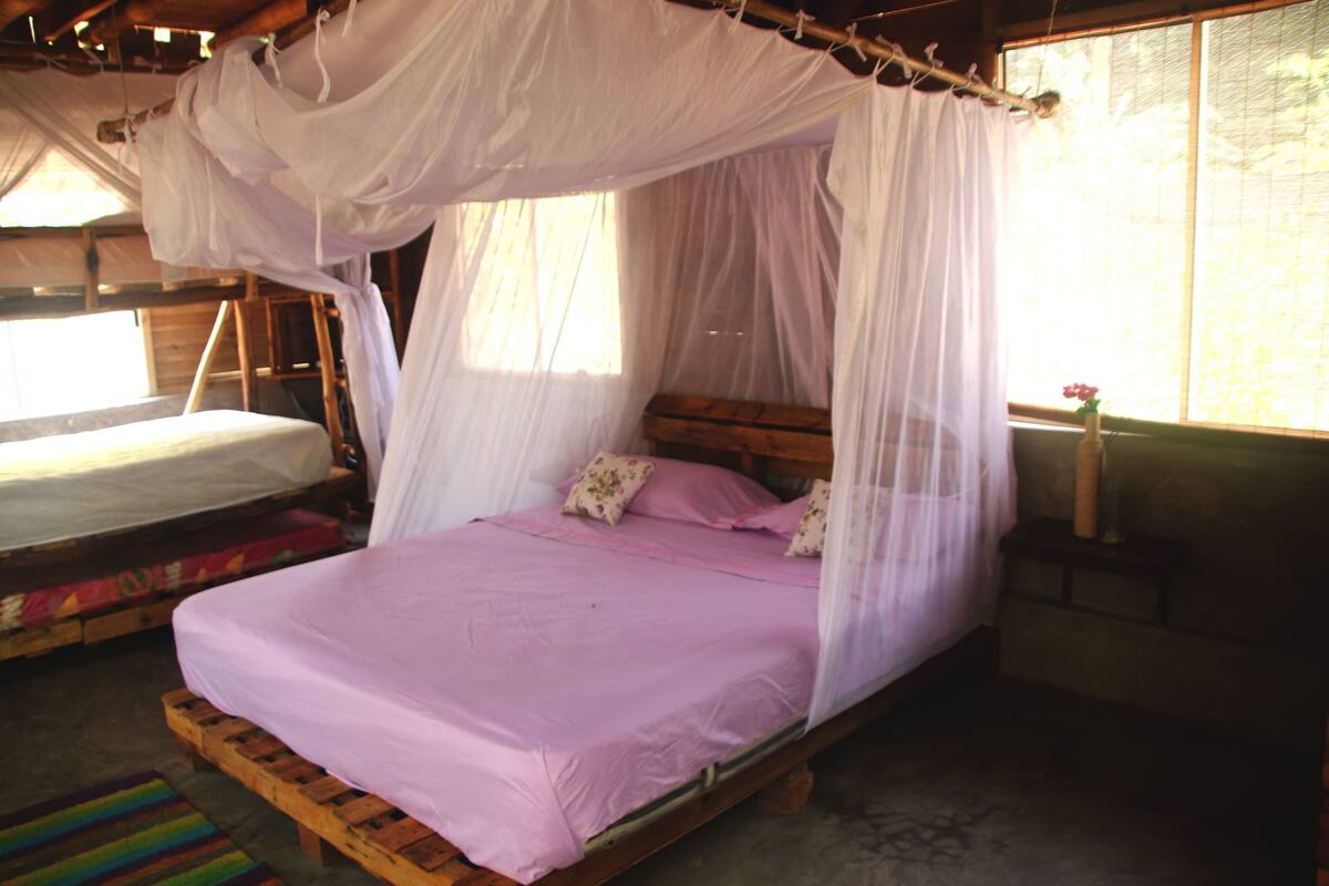 A room with a bed under a canopy, a second bed, a wooden bench, and a window with a view.
