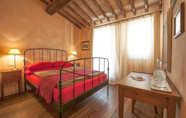 Quarto com estrutura de cama de metal, mesa de cabeceira de madeira com abajur, mesa de madeira com garrafa de vidro e janela com cortinas.