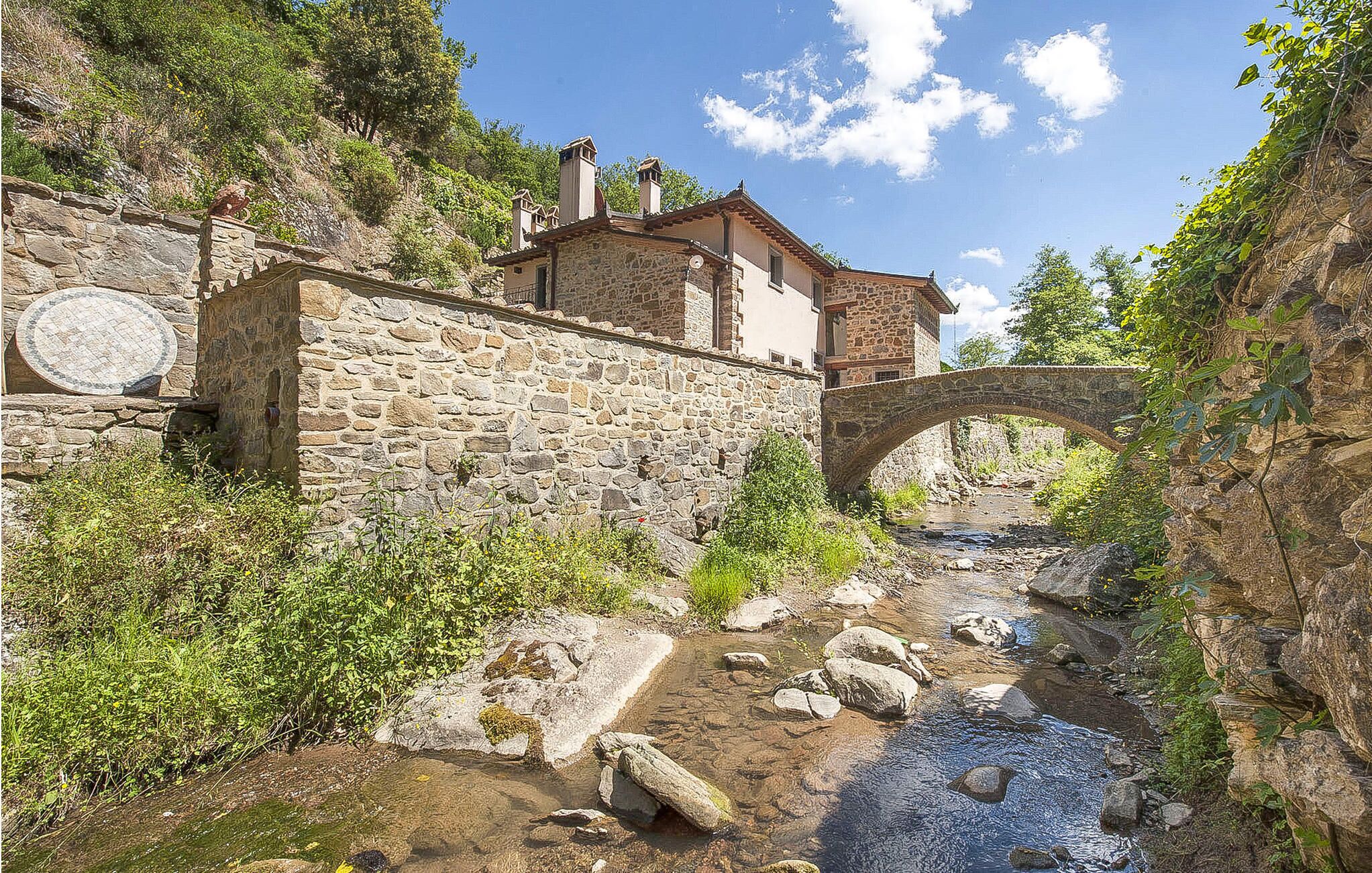 Un bâtiment en pierre, surmonté d’un pont de pierre qui enjambe un ruisseau, le tout entouré de verdure.
