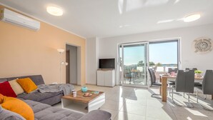A modern living room with a grey sofa, a coffee table, and a TV stand. There is a balcony with a dining area and a view of the sea.