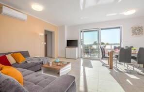 Una sala de estar moderna con un sofá gris, una mesa de centro y un mueble para televisión. Hay un balcón con comedor y vistas al mar.