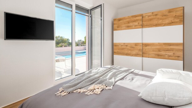 A modern bedroom with a large window, a TV, a wooden headboard, and a bed with white pillows.