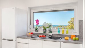 A kitchen with a large window, a sink, a refrigerator, and various fruits and vegetables on the counter.