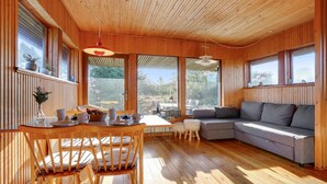 A cozy cabin interior with a wooden dining table, chairs, a sofa, and large windows offering a view of the outdoors.