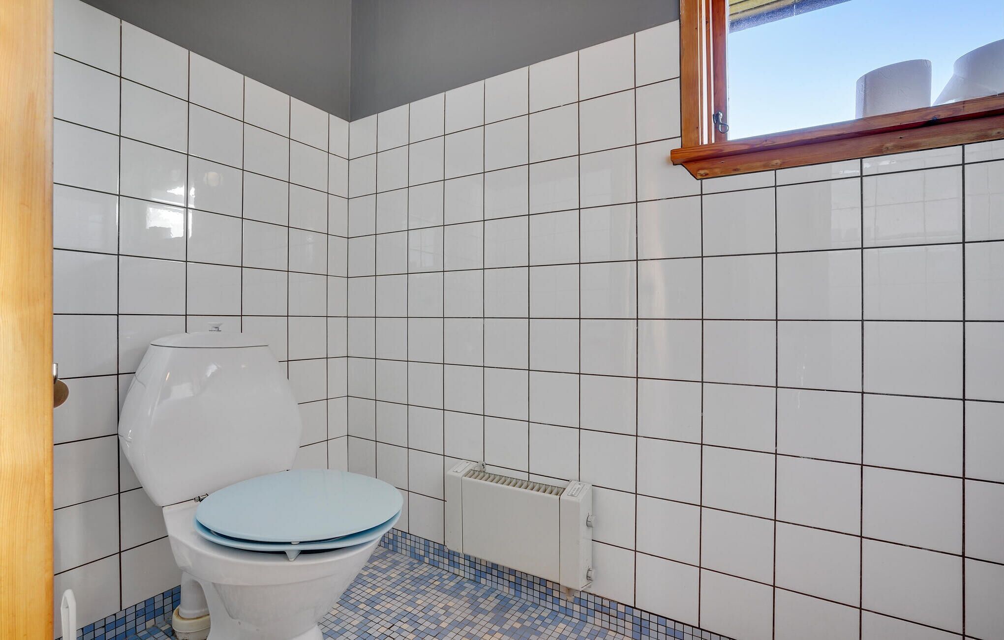 Een kleine betegelde badkamer met een wit toilet, een blauwe tegelvloer en een raam boven het toilet.