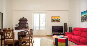 Un coin repas avec une table et des chaises en bois, un téléviseur, un canapé rouge et un pouf coloré.