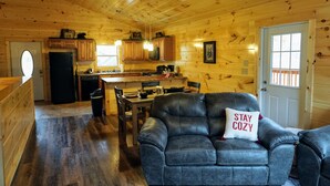 Interior acogedor de una cabaña con comedor, cocina y un sofá de cuero con un cojín que dice 'STAY COZY'.