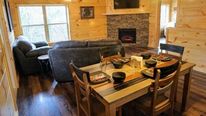 Interior acogedor de una cabaña con comedor, sofá, chimenea y televisor sobre la chimenea.