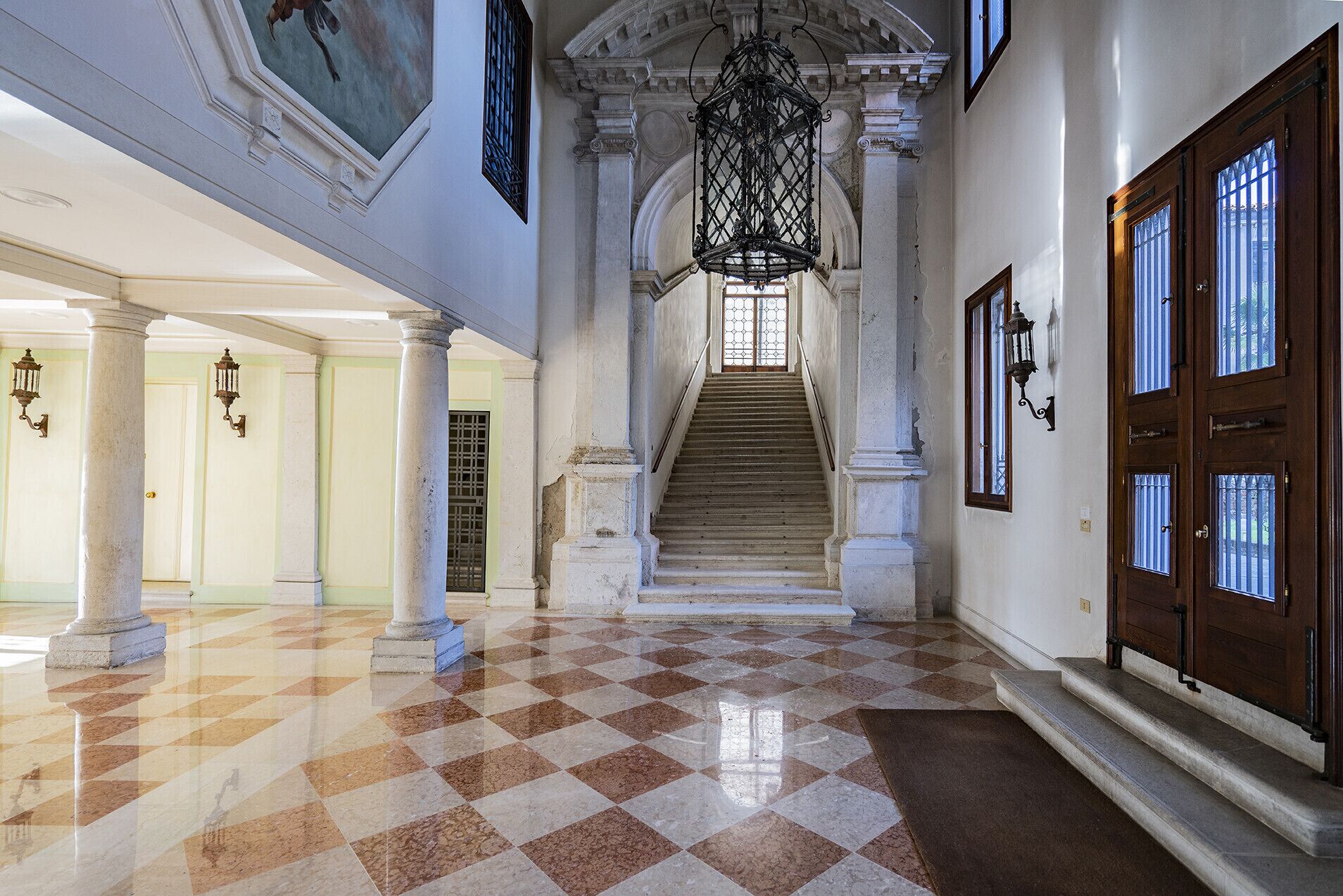 A grand hallway with a checkered floor, a staircase, and a large chandelier.