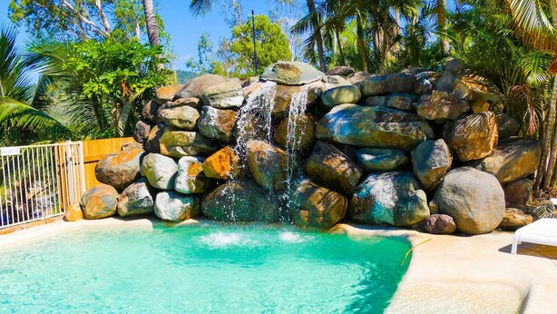 A pool with a waterfall feature surrounded by large rocks and palm trees. - Spa Haven Deluxe 17A (Airlie Beach)