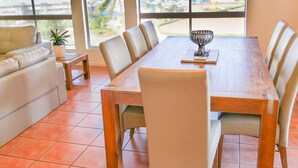 A dining area with a wooden table and chairs, a potted plant, and a view of the beach and marina through large windows. - Spa Haven Deluxe 17A (Airlie Beach)