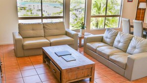 A living room with two beige sofas, a wooden coffee table, and large windows with a view of a beach and palm trees. - Spa Haven Deluxe 17A (Airlie Beach)