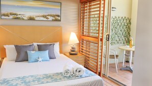 A hotel room with a bed, a nightstand with a lamp, a window with a view of the beach, and a small table with a chair. - Spa Haven 17B (Airlie Beach)